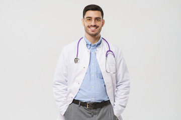 Smiling young male doctor with stethoscope around neck, wearing white coat and round glasses standing isolated on gray background