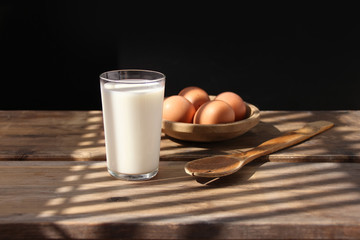 brown eggs lie on a wooden bowl, an old spoon and a glass of white milk in the rays of light on the table, retro concept