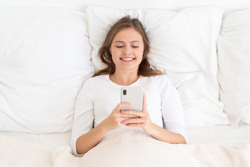 Obraz na płótnie Canvas Young female looking at phone screen while lying in bed in morning, smiling positively, satisfied with good news from friends