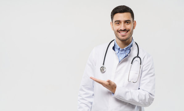 Horizontal Banner Of Smiling Young Male Doctor Showing And Presenting Something With Hand, Isolated On Gray Background With Copy Space On The Left Side