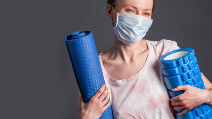 Young caucasian white woman wearing protective mask and holding yoga mat. Quarantine time with pleasure concept. Self isolation, quarantine due to coronavirus