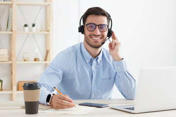 Busy young male working in office, sitting at desk with laptop and takeaway coffee, wearing glasses...