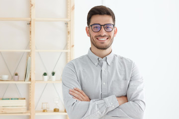 Portrait of young business man wearing gray shirt and glasses, stand with crossed arms in office,...