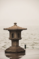 A traditional Chinese stone lantern stands on the edge of West Lake in Hangzhou. The background is blurry in the fog.