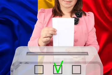 female voter lowers the ballot in a transparent ballot box against the background of the national flag of Romania, concept of state elections, referendum