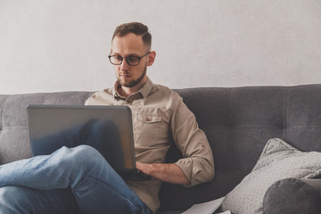 Caucasian young man freelancer working from home. Sitting with laptop on sofa. Surfing internet...