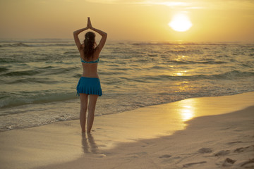 Yoga pose. Woman standing on the beach, practicing yoga. Young woman raising arms with namaste mudra during sunset golden hour. View from back. Melasti beach, Bali.