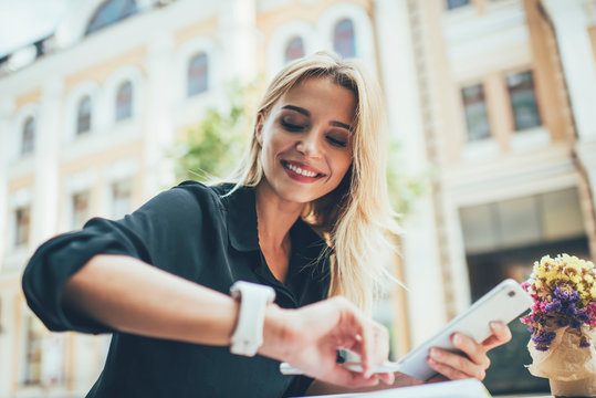 Cheerful woman checking time on modern smartwatch during web networking via cellphone gadget, happy Caucasian female blogger 20 years old using synchronized technology during online browsing in city