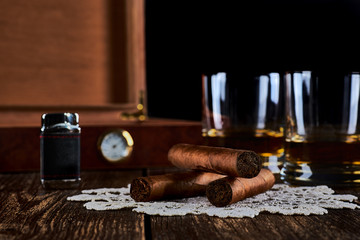 Still life with three cuban cigars, two glasses of whiskey or rum, lighter and wooden box with hygrometer. Old wooden table top and black background.