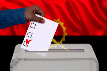 male voter drops a ballot in a transparent ballot box against the background of the Angola national flag, concept of state elections, referendum