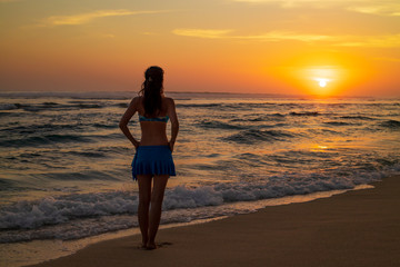 Fototapeta premium Young Caucasian woman walking along the beach. Sunset golden hour. Travel concept. View from back. Copy space. Melasti beach, Bali, Indonesia.