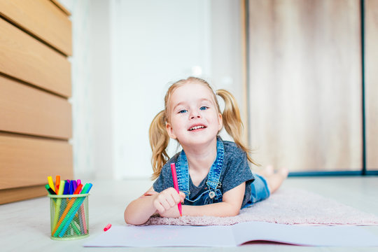 Blonde happy little girl with two ponytales drawing and writing lying on the floor at home. Preschool education, early learning