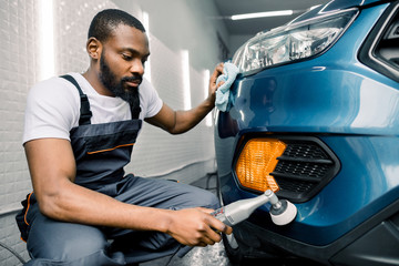 Car detailing series, polishing concept. Professional African male auto service worker, wearing white t-shirt and gray overalls, waxing and polishing blue car with polish machine