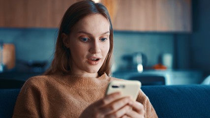 Beautiful surprised caucasian woman in beige sweater sitting on a blue sofa and using smartphone or cell phone in modern room. Tapping, scrolling, watching video, content, bloggs.