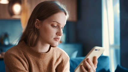 Close up view of cute redhair young woman sitting of the sofa at home using phone. Communication female looking message at cell phone or smartphone.