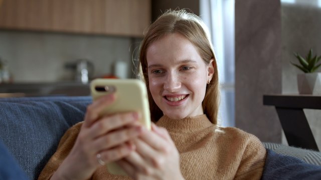 Pretty Teenager Girl With Blonde Hair Lying On Blue Sofa Or Couch At Home, Using Smartphone Or Cell Phone, Watching Something Ang Smiling. Close Up