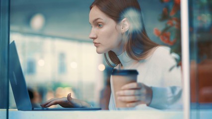 Close up view through the window of attractive business young woman using laptop, checking email, working and drinking some coffee in cafe