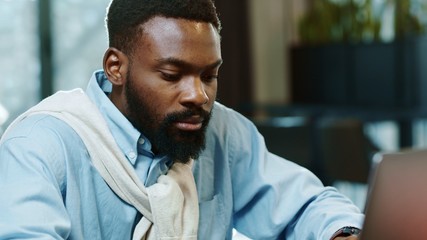 Obraz premium Portrait of african american handsome male using laptop for work in modern office programmer serious looks at monitor screen freelance successful worker businessman close up