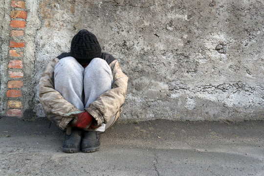 Dirty homeless woman sits under a gray wall