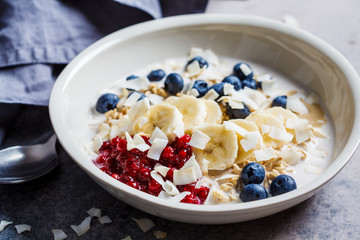 Overnight oatmeal with berries, banana, jam and coconut in a white bowl, gray background.
