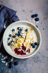 Overnight oatmeal with berries, banana, jam and coconut in a white bowl, top view, gray background.