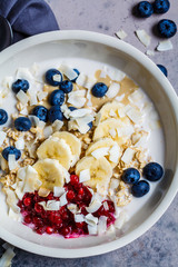 Overnight oatmeal with berries, banana, jam and coconut in white bowl, top view, gray background.