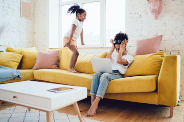 Black daughter jumping on sofa near exhausted mother