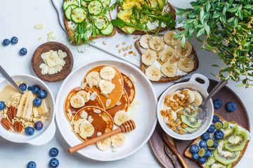 Flat lay of healthy vegetarian breakfast on white background. Oatmeal with fruits, chia pudding, pancakes and toasts with fruits and vegetables.