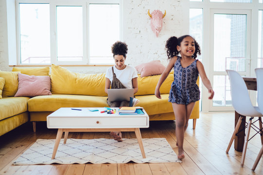 Black Woman Working On Laptop With Daughter Running Around