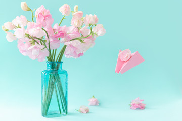 Obraz premium Bouquet of pink sweet pea Lathyrus in beautiful transparent vase on blue background.Paper airplane with message. Spring concept. International Women's Day, March 8 and Valentine's Day,14 of February