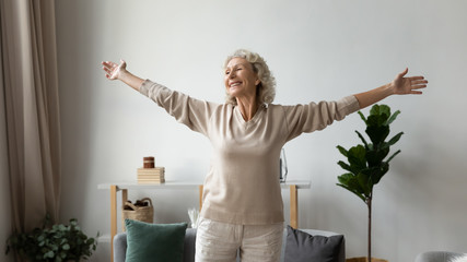 Excited elderly mature retired woman dancing in living room with widely opened outstretched arms,...