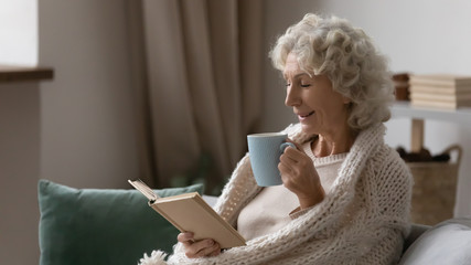 Side view joyful mature older woman covered in knitted plaid reading paper book with poetry,...