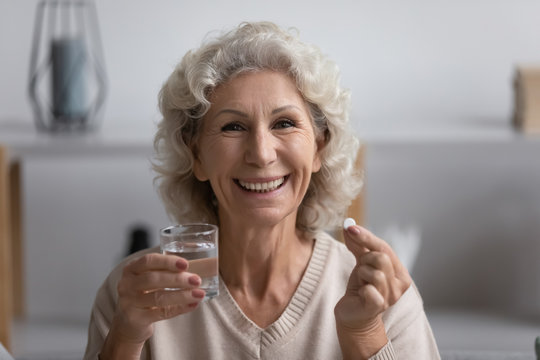 Head Shot Close Up Portrait Happy Elderly Senior Woman Looking At Camera, Holding Glass Of Fresh Water And Pill. Smiling Mature Grandmother Taking Vitamins, Enjoying Daily Healthcare Habit At Home