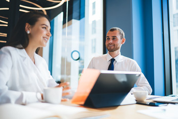 Cheerful male and female colleagues enjoying friendly brainstorming during working time in office, happy man and woman sitting at desk and smiling while discussing business ideas for project