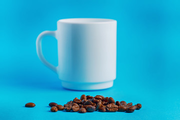 heap of coffee beans in front of a white washed cup on a blue background