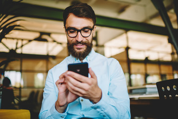 Caucasian hipster guy in trendy spectacles using 4g wireless for web networking via cellphone technology, youthful bearded man typing text content for sharing to social page via modern mobile phone