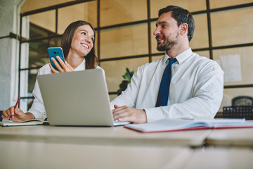 Happy male and female colleagues sitting at table desktop with modern cellphone and laptop devices and communicating about brainstorming cooperation during work day, concept of business collaboration