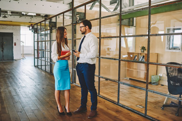 Side view of smiling cute female and confident male in stylish formal clothing discussing creative ideas while standing in office, successful employees talking about collaboration brainstorming
