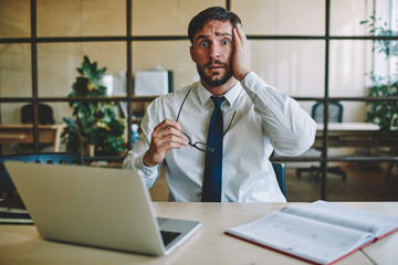 Portrait of bearded Caucasian marketer dressed in formal wear wondering with deadline tasks, surprised male employee holding glasses looking at camera while sitting at table desk in office interior