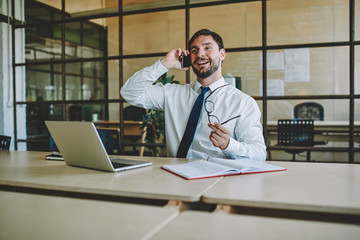 Obraz premium Successful cheerful entrepreneur having mobile conversation with partner sitting at desk in office connecting to 4g internet, happy businessman talking on modern smartphone and smiling indoors