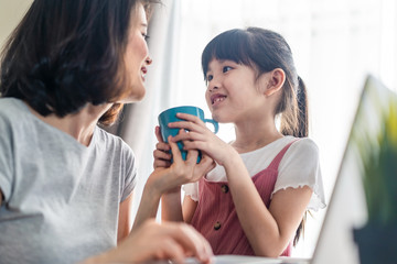 Asian young office mother work from home during coranavirus outbreak. Cute daughter give a cup of coffee to mom and kiss for a break and rest. Love care in family. Covid-19, Social distancing concept