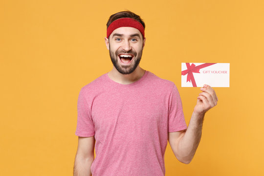 Excited young bearded fitness sporty guy 20s sportsman in headband t-shirt spend weekend in home gym isolated on yellow background. Workout sport motivation lifestyle concept. Hold gift certificate.