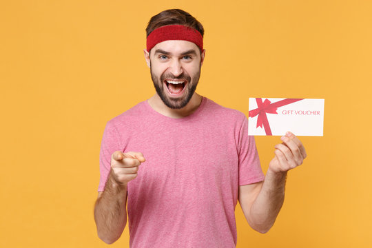 Excited bearded fitness sporty guy sportsman in headband t-shirt in home gym isolated on yellow background. Workout sport motivation concept. Hold gift certificate, pointing index finger on camera.