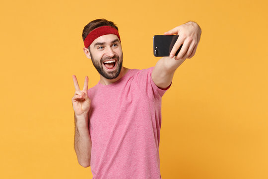 Excited bearded fitness sporty guy sportsman in headband t-shirt in home gym isolated on yellow background. Workout sport motivation concept. Doing selfie shot on mobile phone, showing victory sign.