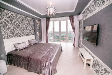 A grey bedroom with a bed and a large panoramic window to the floor. With chandelier, TV and bedside table