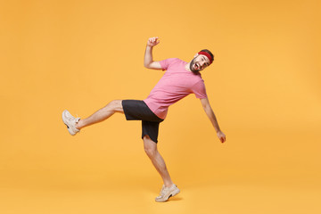 Fototapeta na wymiar Side view of cheerful young bearded fitness sporty guy sportsman in headband t-shirt in home gym isolated on yellow background. Workout sport motivation lifestyle concept. Looking camera, rising leg.