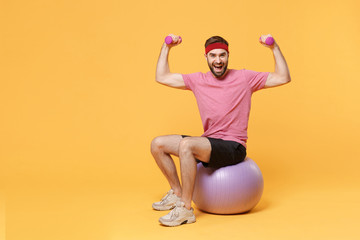 Cheerful bearded fitness sporty guy sportsman in headband t-shirt in home gym isolated on yellow background. Workout sport motivation lifestyle concept. Sit on fitball, doing exercise with dumbbell.
