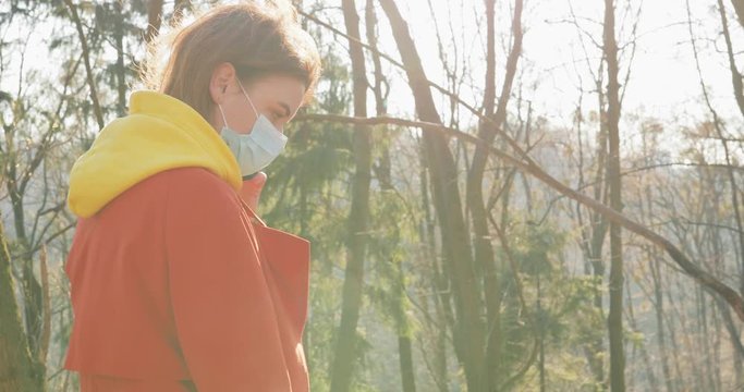 COVID-19 coronavirus infection Woman dressed in orange red coat wearing Protective Face Mask walking in the park with dog. Sunny day Sun flayers pandemic disease virus epidemic air.