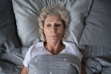 Head shot top above view stressed middle aged retired woman lying in bed under duvet with opened...