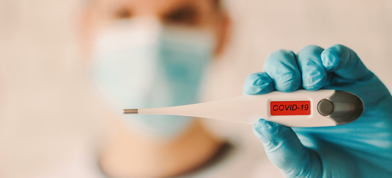 Closeup man in medical face mask and glove showing digital thermometer with COVID-19 text. Doctor in protective glove and mask holding electronic thermometer in hand. Coronavirus symptoms, diagnosis